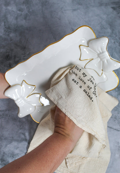 A person dries a white ceramic plate with bow details using the “I’m pretty sure being friends...” Tea Towel by Hill Country Chocolate—a beige towel made from unbleached cotton with text printed on it. This natural kitchen towel adds both charm and practicality to the kitchen.