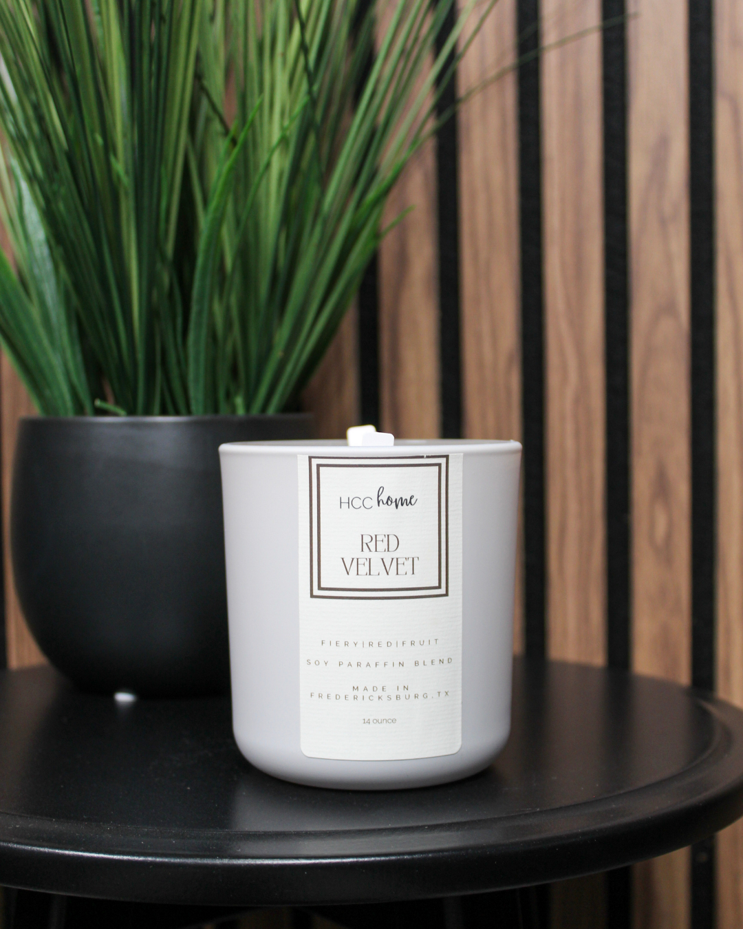 A hand-poured white candle, labeled “HCC Home Candle - Red Velvet” by Hill Country Chocolate, sits on a black table next to a black pot with tall green grass-like plants. A wood-paneled wall is in the background.