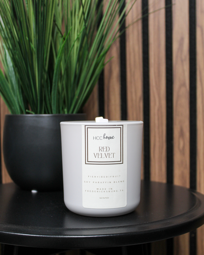 A hand-poured white candle, labeled “HCC Home Candle - Red Velvet” by Hill Country Chocolate, sits on a black table next to a black pot with tall green grass-like plants. A wood-paneled wall is in the background.