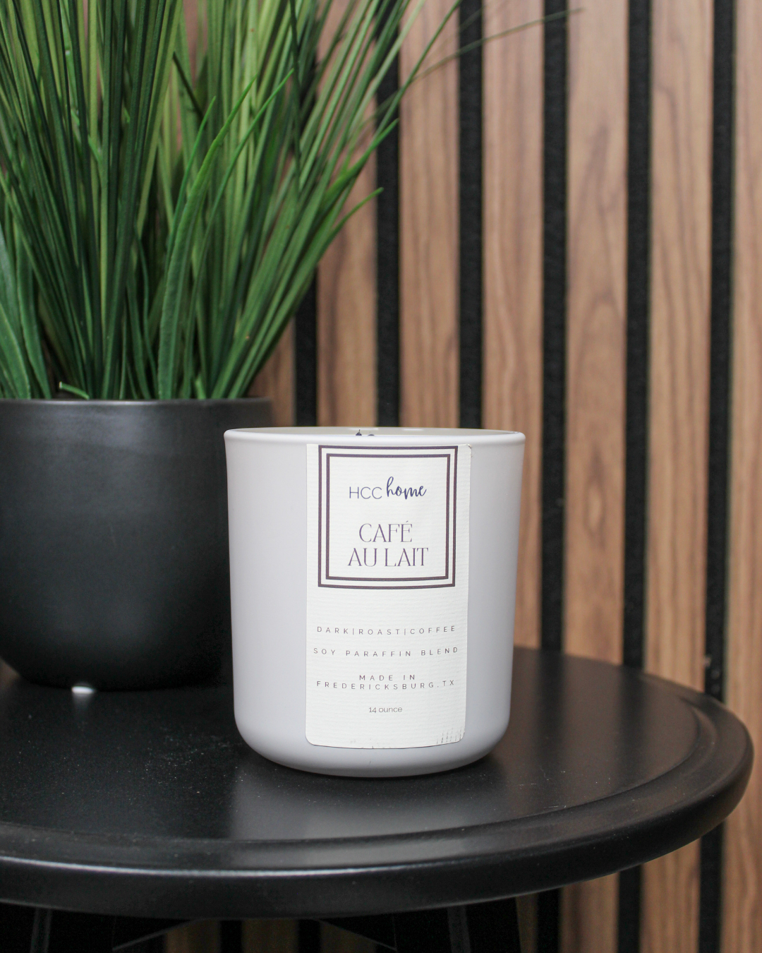 A hand-poured HCC Home Candle - Cafe Au Lait by Hill Country Chocolate sits on a black round table next to a black pot of green, grass-like plants, set against a backdrop of wood panels and black slats—an inviting Texas-made candle perfect for creating cozy spaces.