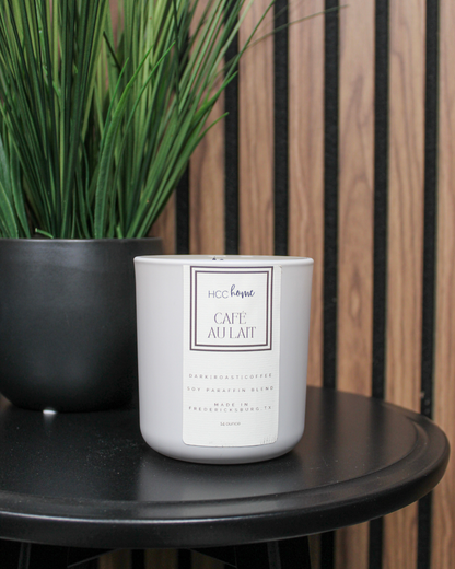 A hand-poured HCC Home Candle - Cafe Au Lait by Hill Country Chocolate sits on a black round table next to a black pot of green, grass-like plants, set against a backdrop of wood panels and black slats—an inviting Texas-made candle perfect for creating cozy spaces.