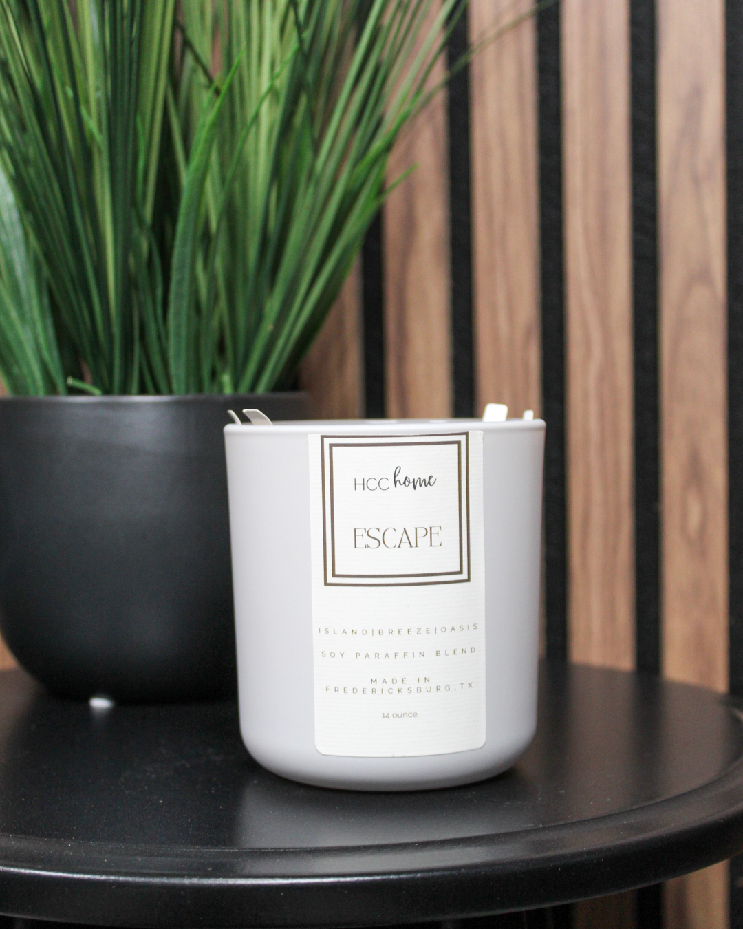 A white candle labeled “ESCAPE” from Hill Country Chocolate’s HCC Home Candle collection sits on a black table beside a black pot with tall green grass-like plants. Hand poured in Fredericksburg, this luxurious scent candle is a distinctive Texas product.