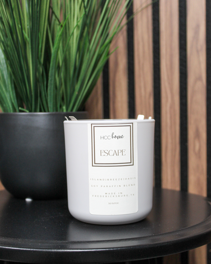 A white candle labeled “ESCAPE” from Hill Country Chocolate’s HCC Home Candle collection sits on a black table beside a black pot with tall green grass-like plants. Hand poured in Fredericksburg, this luxurious scent candle is a distinctive Texas product.