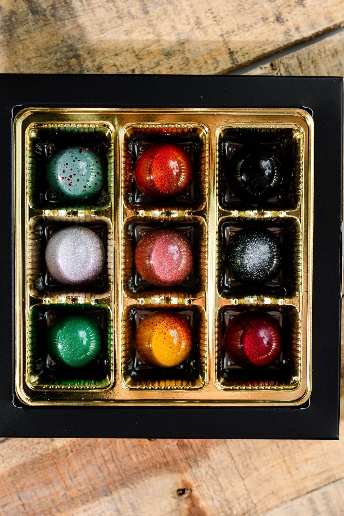 A box of ten assorted, glossy bonbons from the Hill Country Chocolate Artisanal Bonbon Collection, with vibrant colors and patterns, arranged in a gold tray on wood.