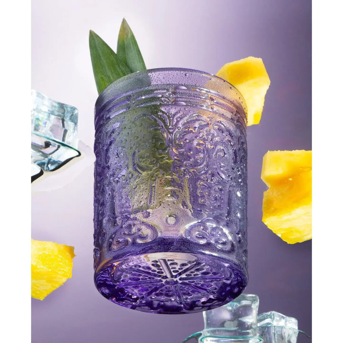 A purple embossed Double Old Fashioned Glass from Hill Country Chocolate—crafted from unleaded crystal and filled with two green leaves—rests amid pineapple pieces on a purple background, promising to elevate any dining experience.