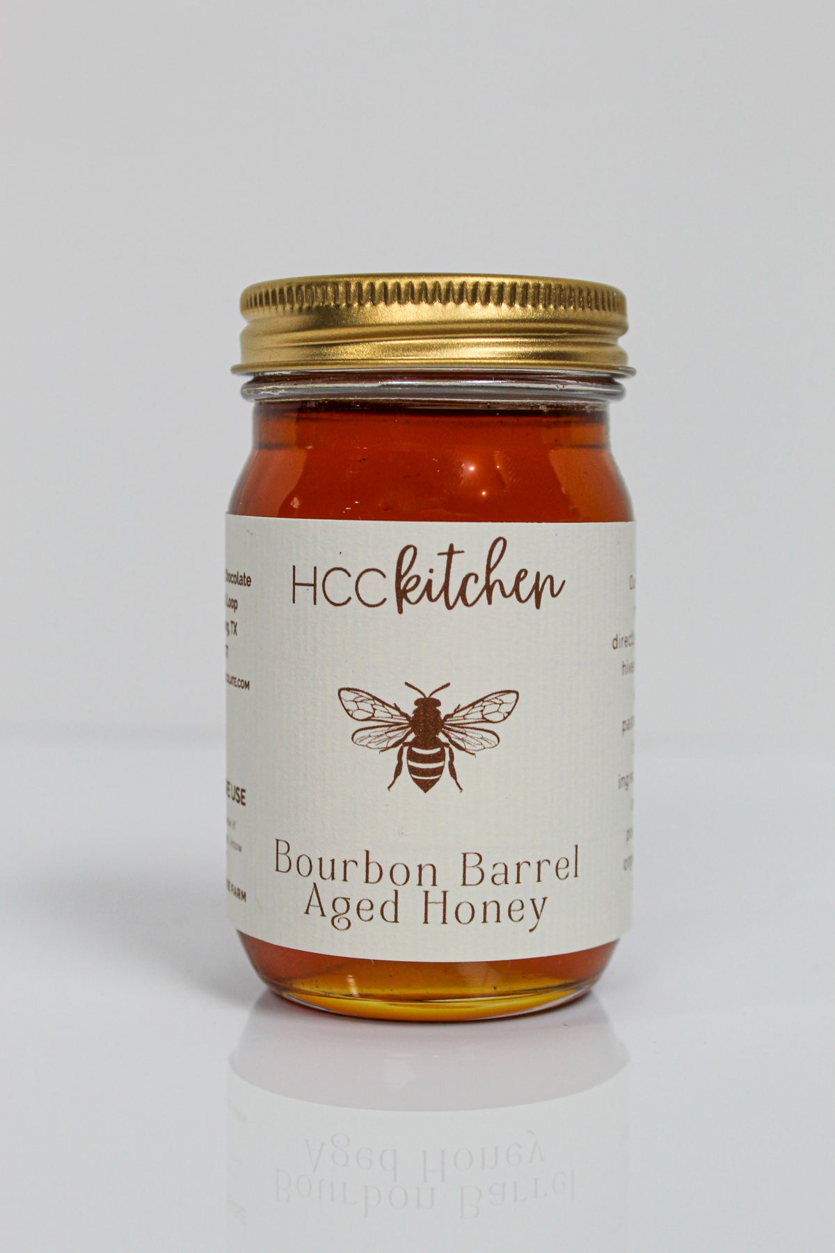 A glass jar with a gold lid labeled “Hill Country Chocolate Bourbon Barrel Aged Honey,” adorned with a bee illustration, delivers a taste of gourmet honey crafted in Texas.