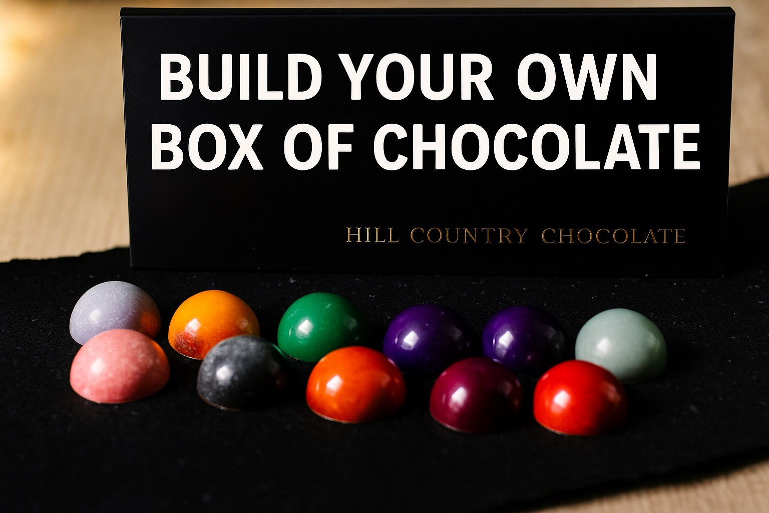 A selection of vibrant artisanal bonbons is showcased in front of a sign saying, "Build Your Own Box of Chocolates" by Hill Country Chocolate.