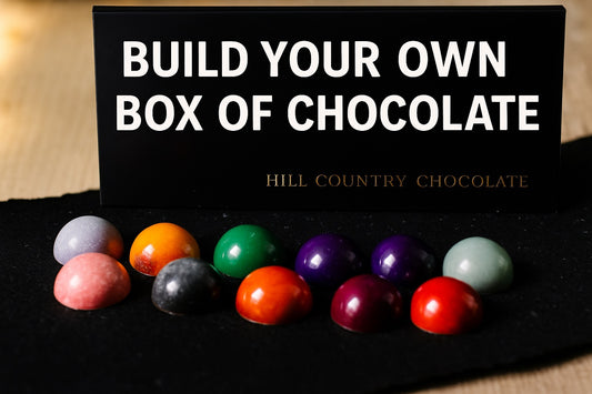 A selection of vibrant artisanal bonbons is showcased in front of a sign saying, "Build Your Own Box of Chocolates" by Hill Country Chocolate.