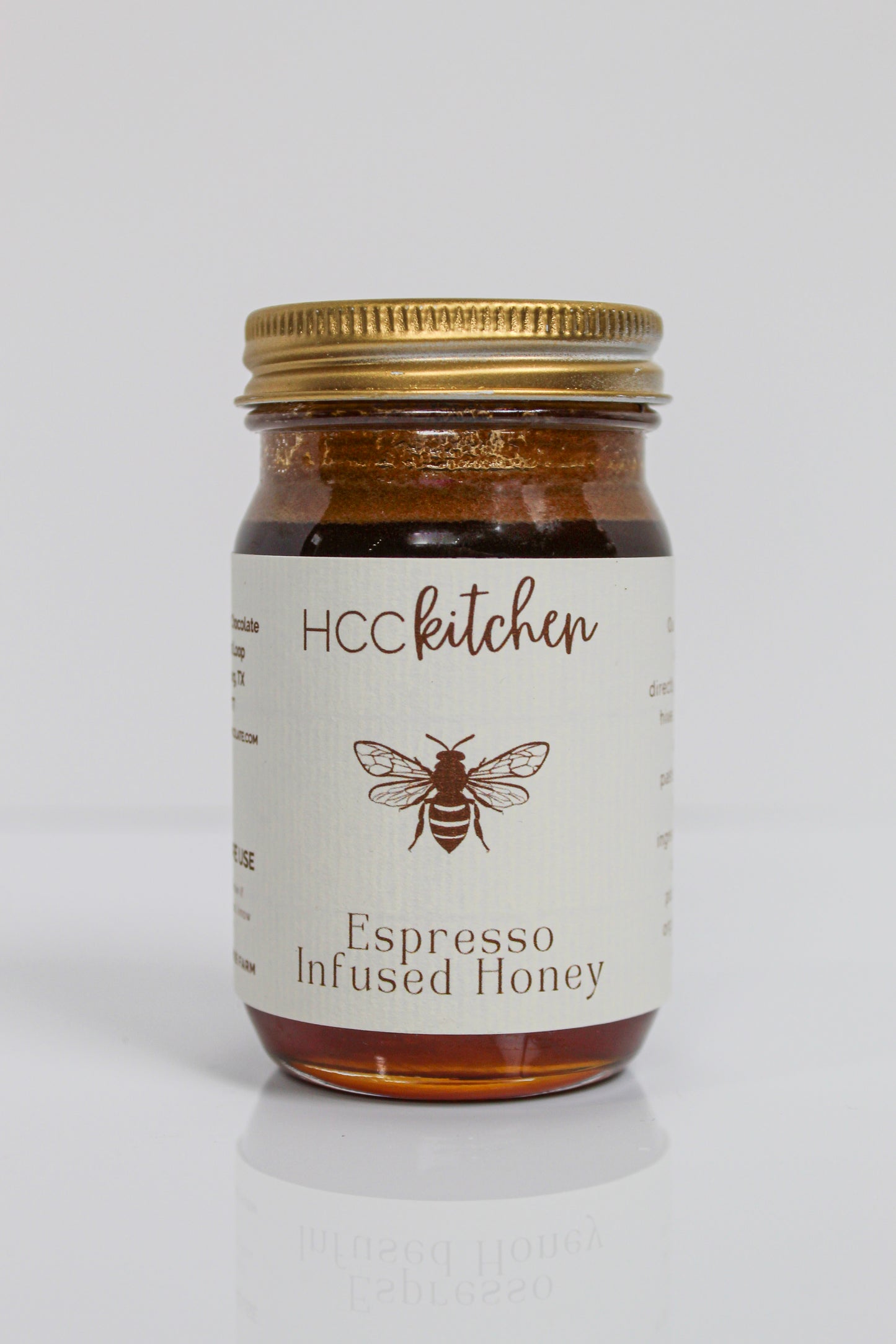 A glass jar labeled “Hill Country Chocolate Espresso Infused Honey,” featuring a gold lid and a bee illustration on the front, displays rich coffee honey made from pure Texas honey, set against a white background.