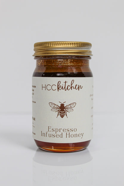 A glass jar labeled “Hill Country Chocolate Espresso Infused Honey,” featuring a gold lid and a bee illustration on the front, displays rich coffee honey made from pure Texas honey, set against a white background.