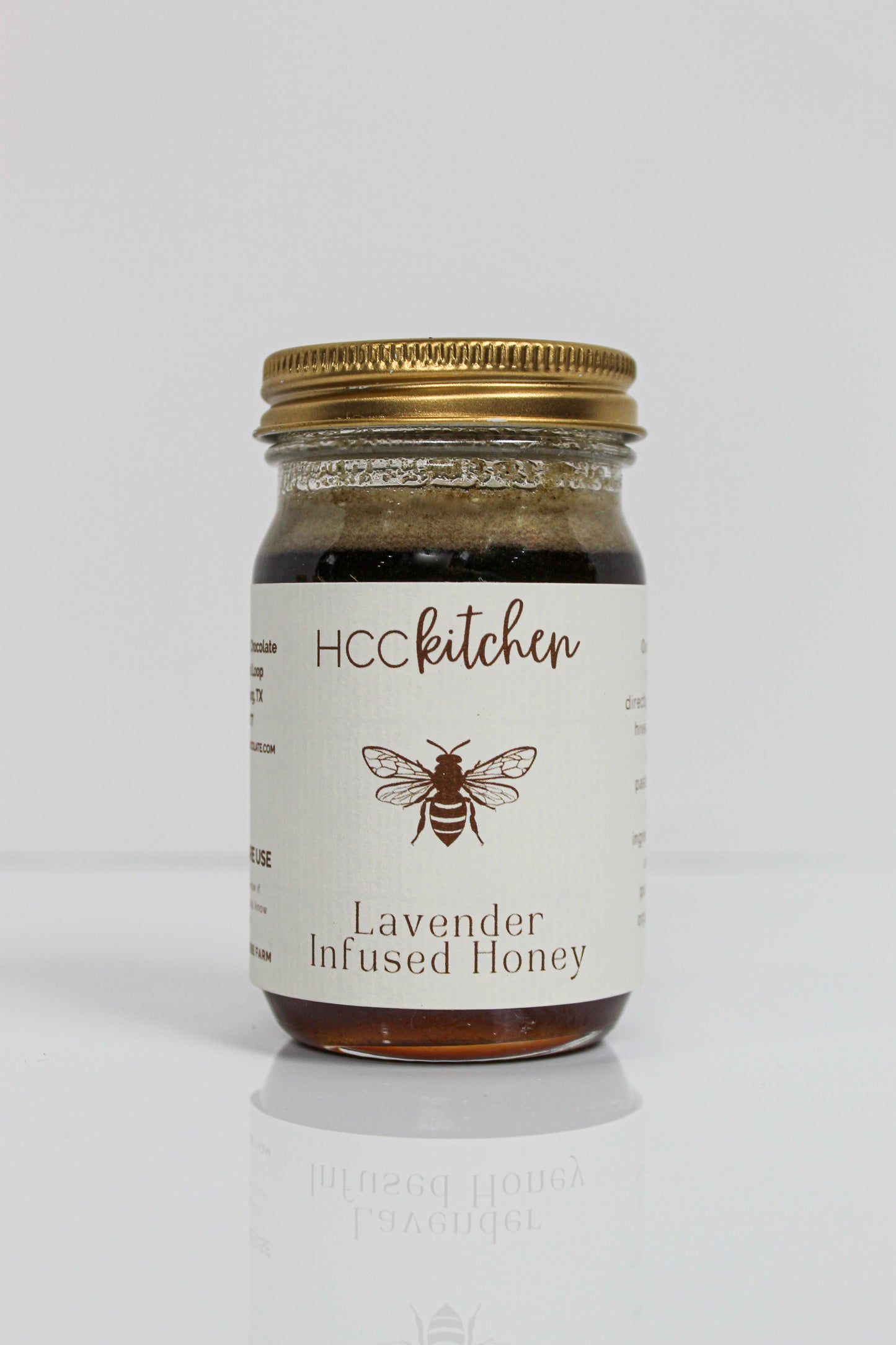 A glass jar labeled “Hill Country Chocolate Lavender Infused Honey” with a gold lid, adorned with a bee illustration on the front, showcases the rich flavors of Texas honey complemented by delicate lavender notes in each spoonful.