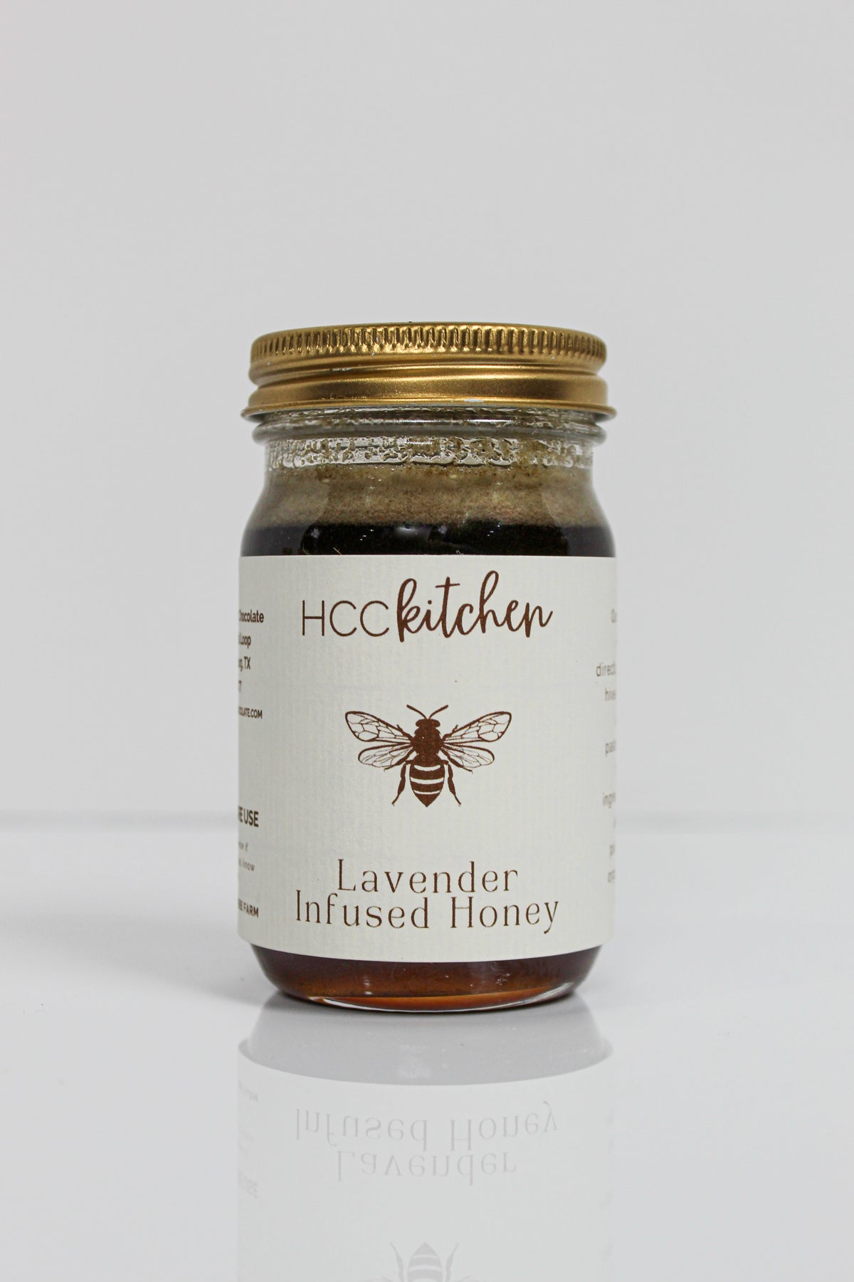 A glass jar labeled “Hill Country Chocolate Lavender Infused Honey” with a gold lid, adorned with a bee illustration on the front, showcases the rich flavors of Texas honey complemented by delicate lavender notes in each spoonful.