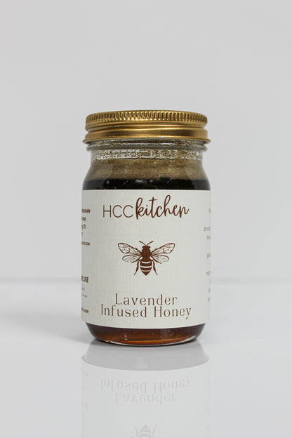A glass jar labeled “Hill Country Chocolate Lavender Infused Honey” with a gold lid, adorned with a bee illustration on the front, showcases the rich flavors of Texas honey complemented by delicate lavender notes in each spoonful.