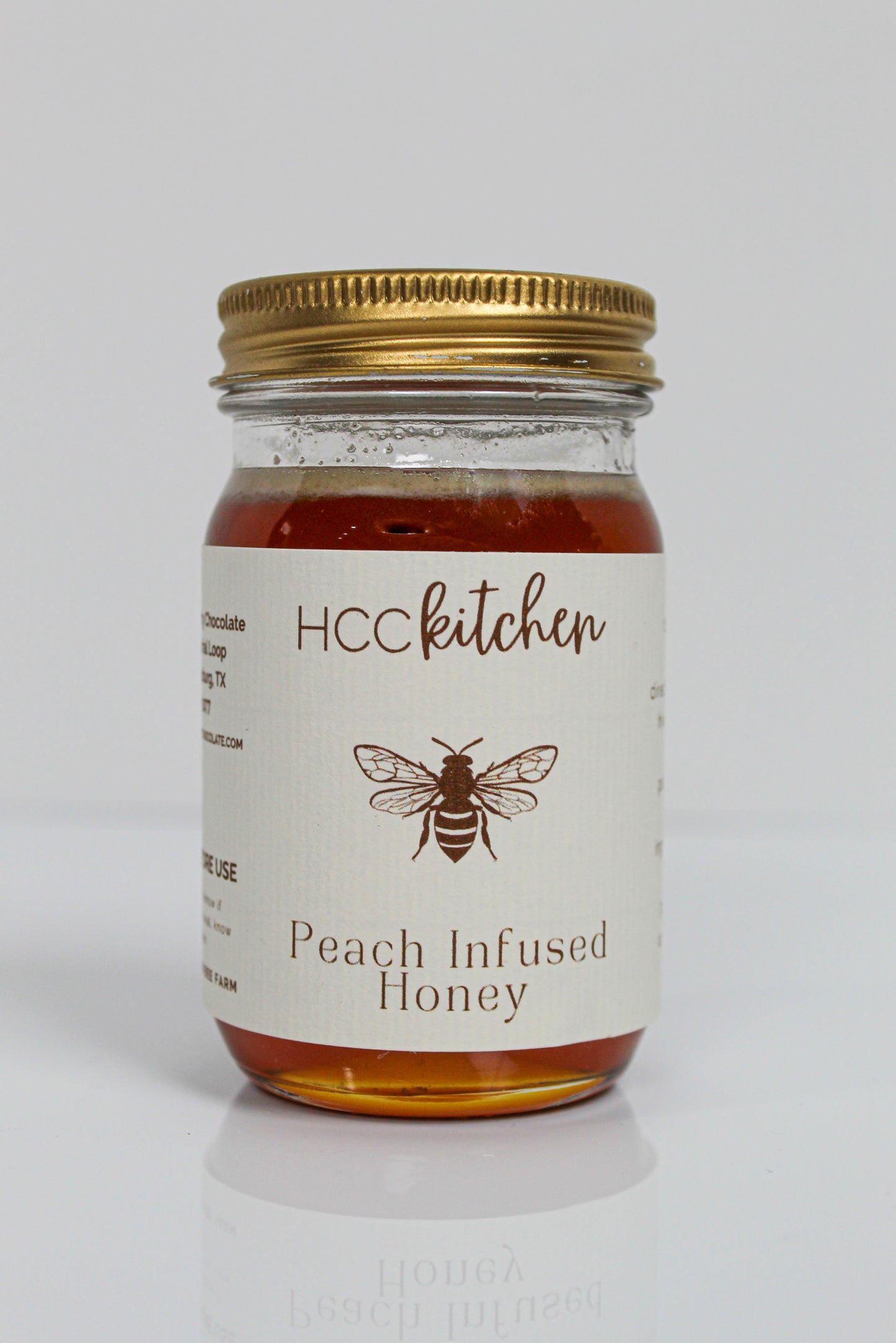 A glass jar of Hill Country Chocolate Peach Infused Honey, made from pure Texas honey, with a gold lid and a white label adorned with a bee illustration.