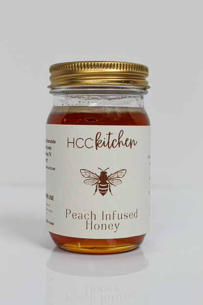 A glass jar of Hill Country Chocolate Peach Infused Honey, made from pure Texas honey, with a gold lid and a white label adorned with a bee illustration.