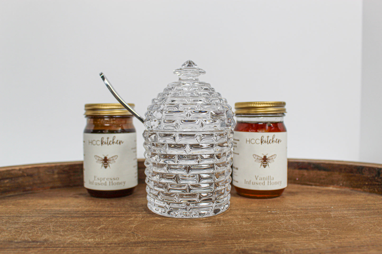 Two jars of Hill Country Chocolate’s Lavender Infused Honey sit on a wooden tray, framing a clear glass honey jar shaped like a beehive in the center and highlighting the rich variety and raw honey freshness in every spoonful.