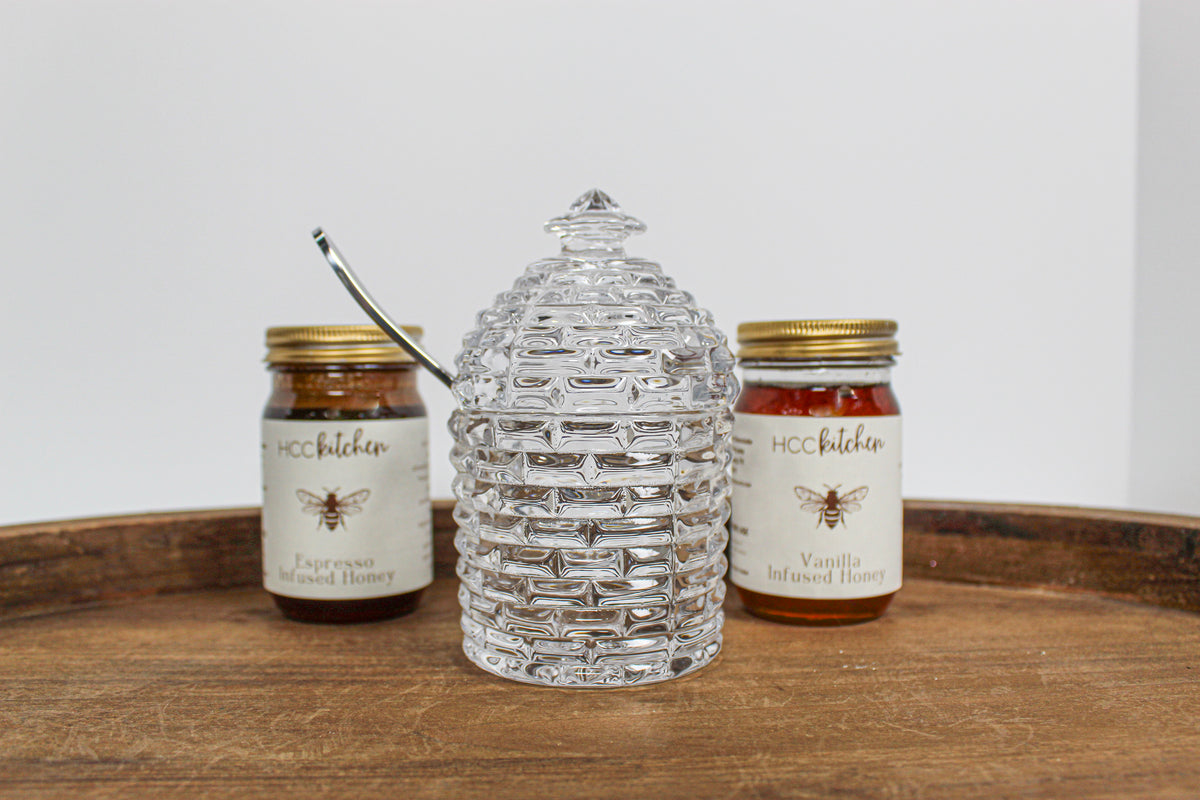 Two jars of Hill Country Chocolate’s Lavender Infused Honey sit on a wooden tray, framing a clear glass honey jar shaped like a beehive in the center and highlighting the rich variety and raw honey freshness in every spoonful.