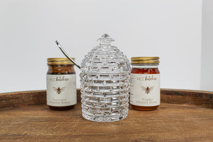 Two jars of Hill Country Chocolate’s Lavender Infused Honey sit on a wooden tray, framing a clear glass honey jar shaped like a beehive in the center and highlighting the rich variety and raw honey freshness in every spoonful.