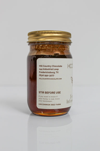 A glass jar with a gold lid contains rich Lavender Infused Honey. The label reads “Hill Country Chocolate” and includes contact details along with instructions to stir before use.