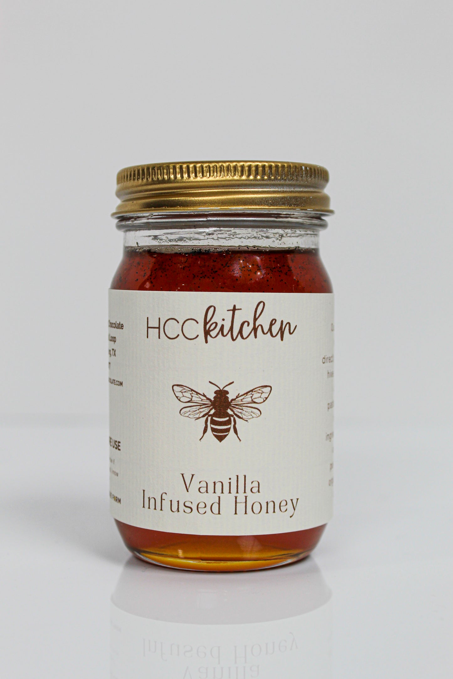 A glass jar with a gold lid labeled “Hill Country Chocolate Vanilla Infused Honey,” containing amber-colored Pure Texas Honey delicately infused with vanilla.