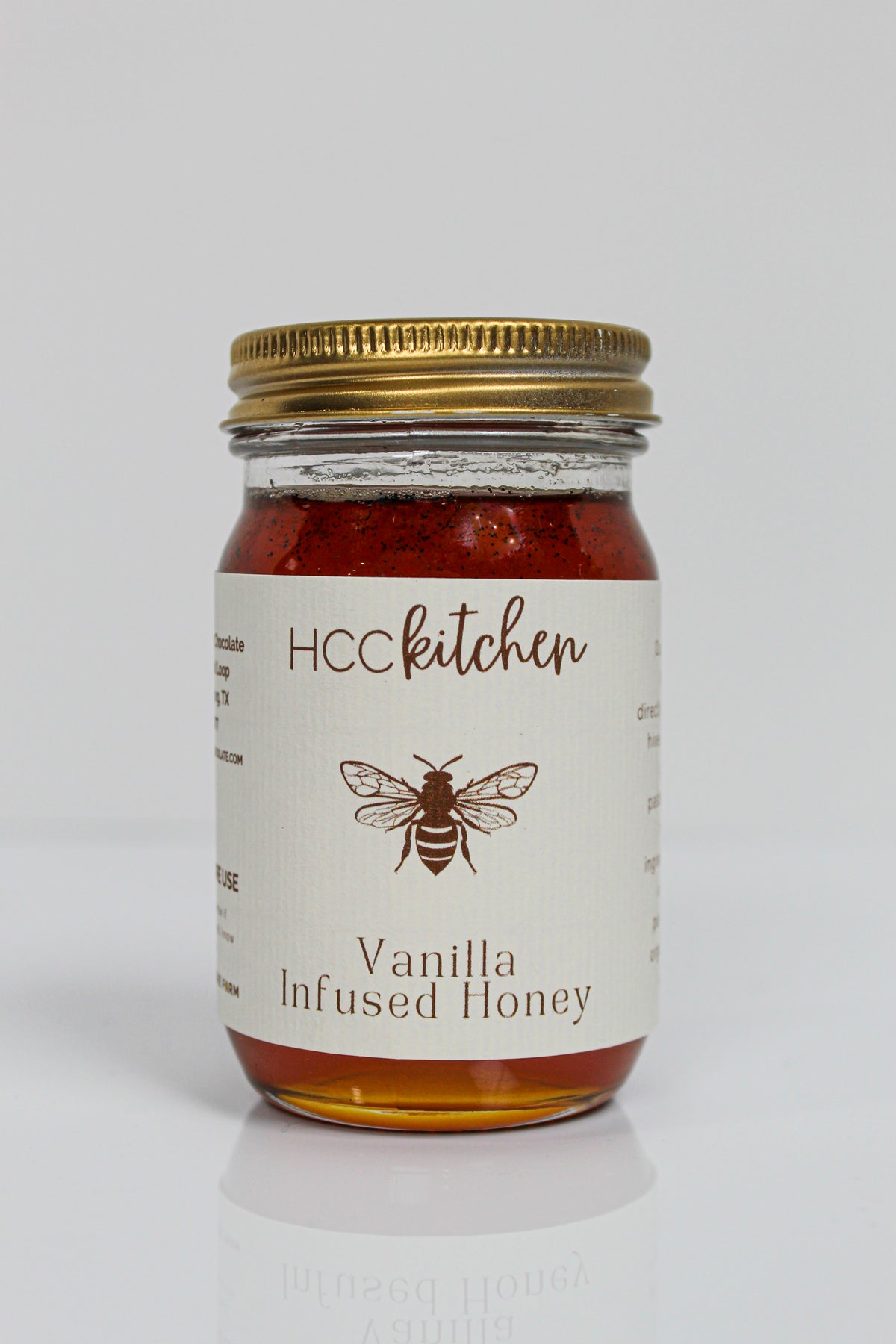 A glass jar with a gold lid labeled “Hill Country Chocolate Vanilla Infused Honey,” containing amber-colored Pure Texas Honey delicately infused with vanilla.