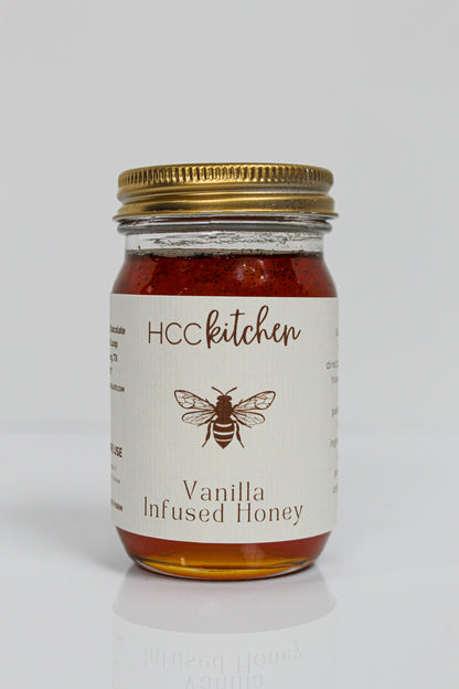 A glass jar with a gold lid labeled “Hill Country Chocolate Vanilla Infused Honey,” containing amber-colored Pure Texas Honey delicately infused with vanilla.