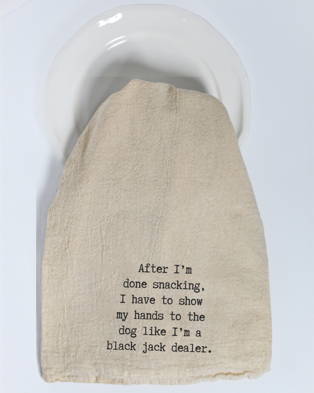 A beige “After I’m done eating...” tea towel by Hill Country Chocolate, featuring black text, covers a white plate. The towel reads: “After I’m done snacking, I have to show my hands to the dog like I’m a black jack dealer.”.