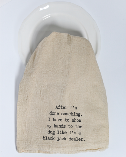 A beige “After I’m done eating...” tea towel by Hill Country Chocolate, featuring black text, covers a white plate. The towel reads: “After I’m done snacking, I have to show my hands to the dog like I’m a black jack dealer.”.