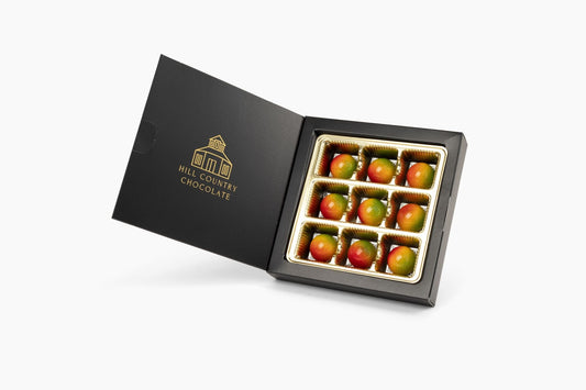 A black box labeled "Hill Country Chocolate" holds nine Fredericksburg Peach Yogurt Bonbons, each glossy and artisanal, arranged neatly in a gold tray.