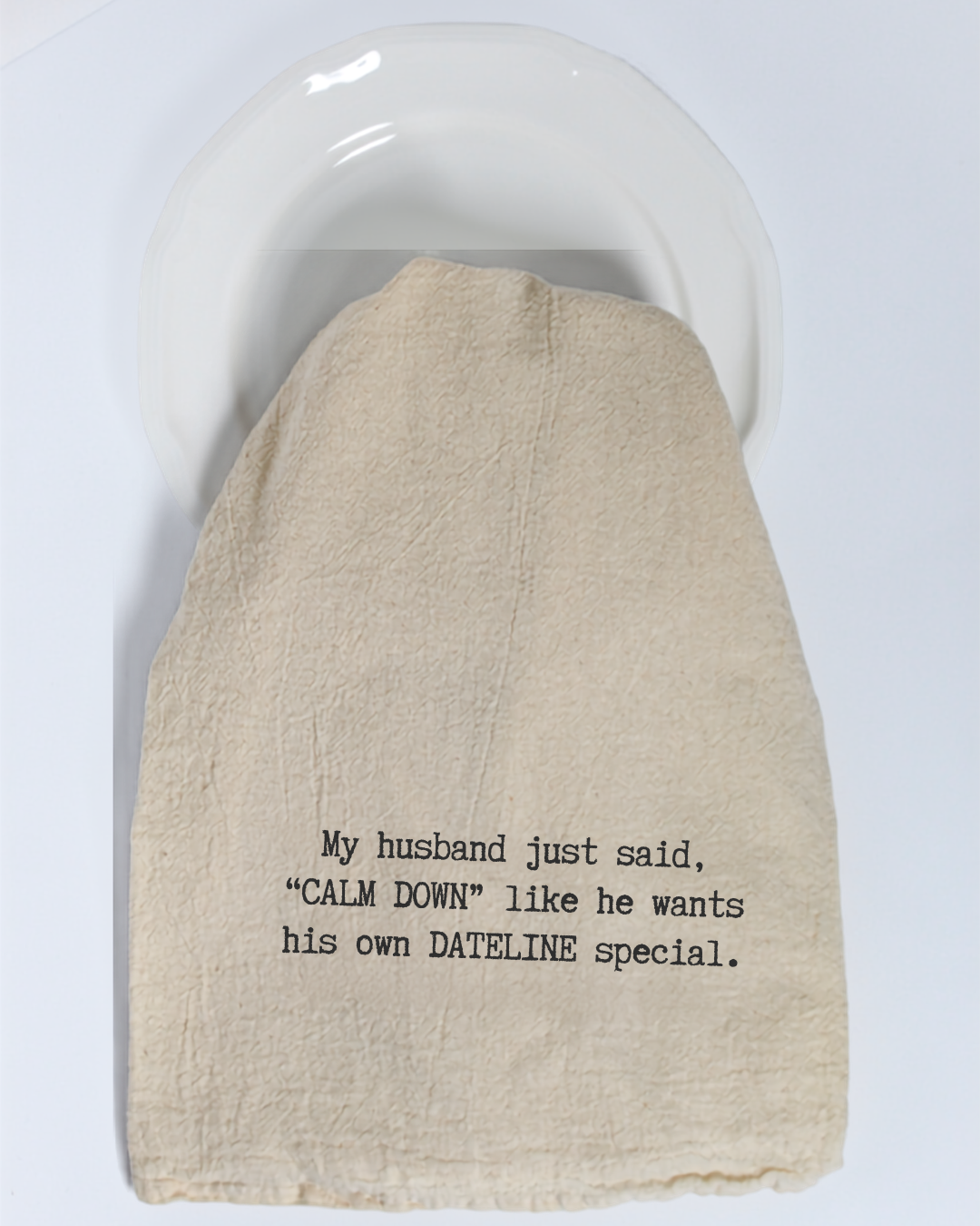 A Hill Country Chocolate “My husband just said...” Tea Towel, featuring eco-friendly beige fabric and black text that reads, “My husband just said, ‘CALM DOWN’ like he wants his own DATELINE special,” is draped over a white plate.
