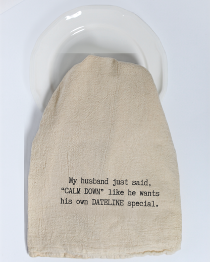 A Hill Country Chocolate “My husband just said...” Tea Towel, featuring eco-friendly beige fabric and black text that reads, “My husband just said, ‘CALM DOWN’ like he wants his own DATELINE special,” is draped over a white plate.
