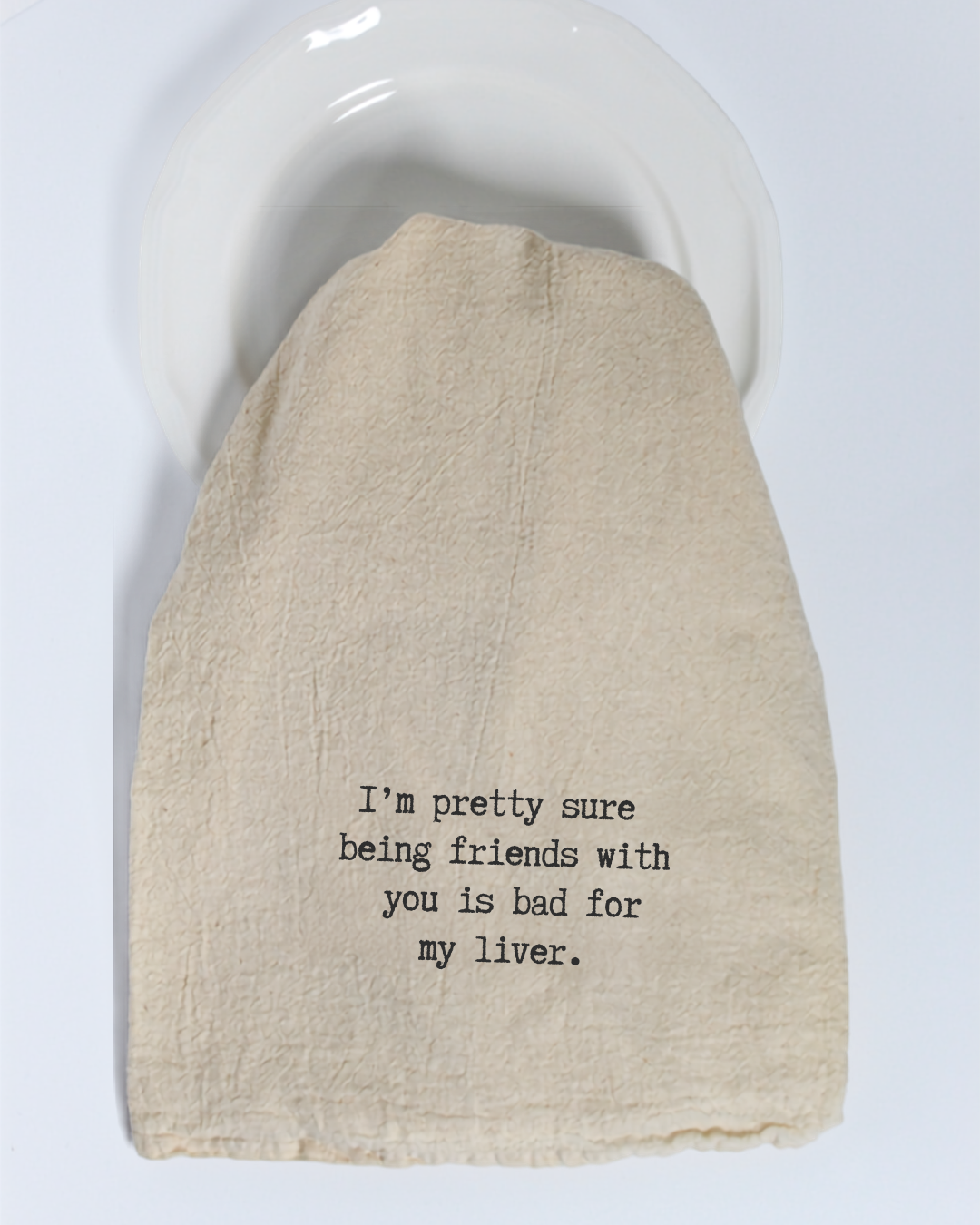 A beige unbleached cotton tea towel by Hill Country Chocolate, featuring the text “I’m pretty sure being friends with you is bad for my liver,” rests on a white plate.