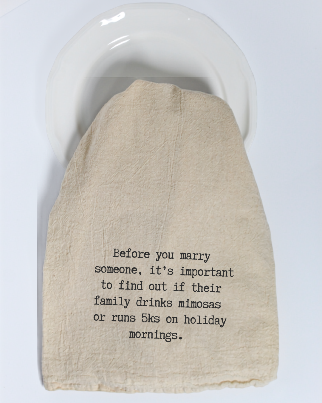 The Hill Country Chocolate “Before you marry someone...” Tea Towel is made of unbleached beige cotton and features black text that reads: “Before you marry someone, it’s important to find out if their family drinks mimosas or runs 5ks on holiday mornings.” Displayed on a white plate, this humorous tea towel is a delightful way to add charm and personality to your kitchen décor.