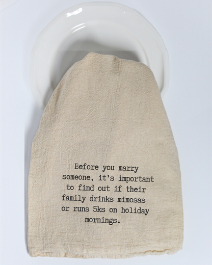 The Hill Country Chocolate “Before you marry someone...” Tea Towel is made of unbleached beige cotton and features black text that reads: “Before you marry someone, it’s important to find out if their family drinks mimosas or runs 5ks on holiday mornings.” Displayed on a white plate, this humorous tea towel is a delightful way to add charm and personality to your kitchen décor.