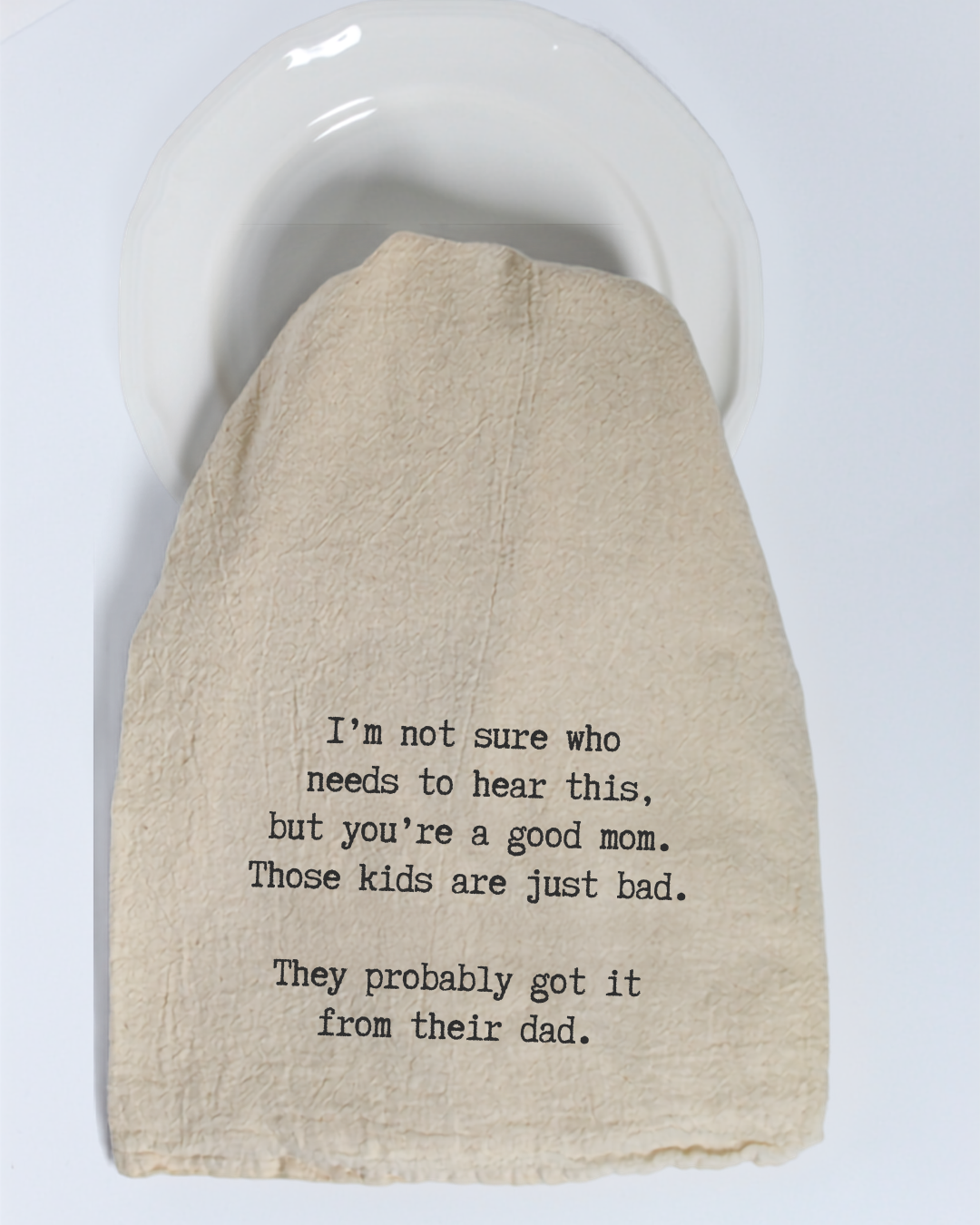 A cream-colored, eco-friendly “I’m not sure who needs to hear this...” tea towel by Hill Country Chocolate, featuring black text that humorously comments on parenting and children’s behavior, is draped over a white plate.