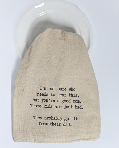 A cream-colored, eco-friendly “I’m not sure who needs to hear this...” tea towel by Hill Country Chocolate, featuring black text that humorously comments on parenting and children’s behavior, is draped over a white plate.