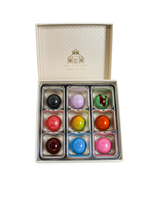 A box of Hill Country Chocolate Artisanal Bonbon Collection containing nine colorful, glossy artisanal bonbons arranged in a three-by-three grid.